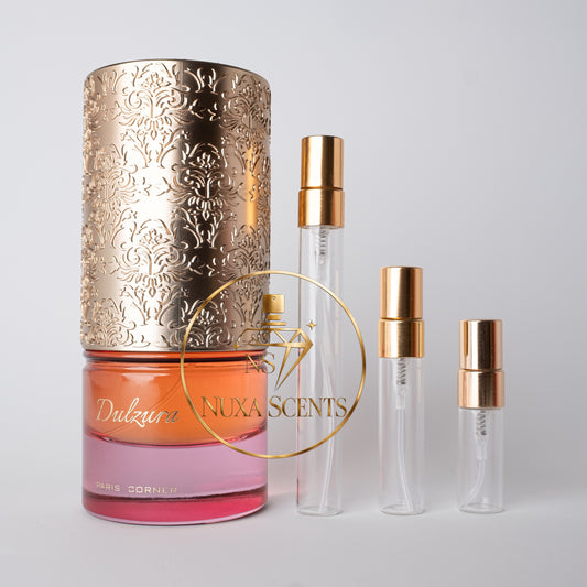 A perfume bottle with a large ornate gold cap and three clear glass travel atomizer vials with gold collars, arranged on a light gray background. A gold circular logo overlay reads 'Dulzura' and 'NUXA SCENTS'.