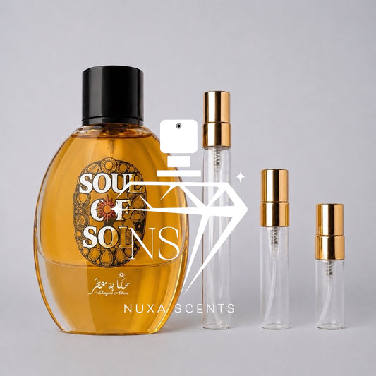 A bottle of Soul of Sol perfume with amber liquid and a black cap, shown alongside three clear glass travel atomizer vials with gold caps on a light gray background. A large white camera-and-diamond watermark with the text 'NUXA SCENTS' overlays the center.